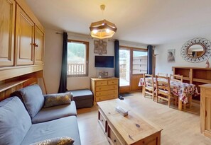Ein gemütliches Wohnzimmer mit einer Couch, einem Holztisch und einem Fernseher. Ein Balkon mit Blick und ein Essbereich mit Holzstühlen und einem Tisch sind ebenfalls vorhanden.