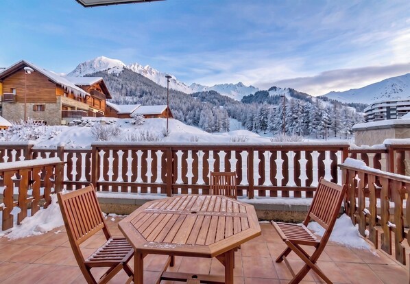 A wooden deck with a table and chairs, overlooking a snowy mountain landscape with buildings.