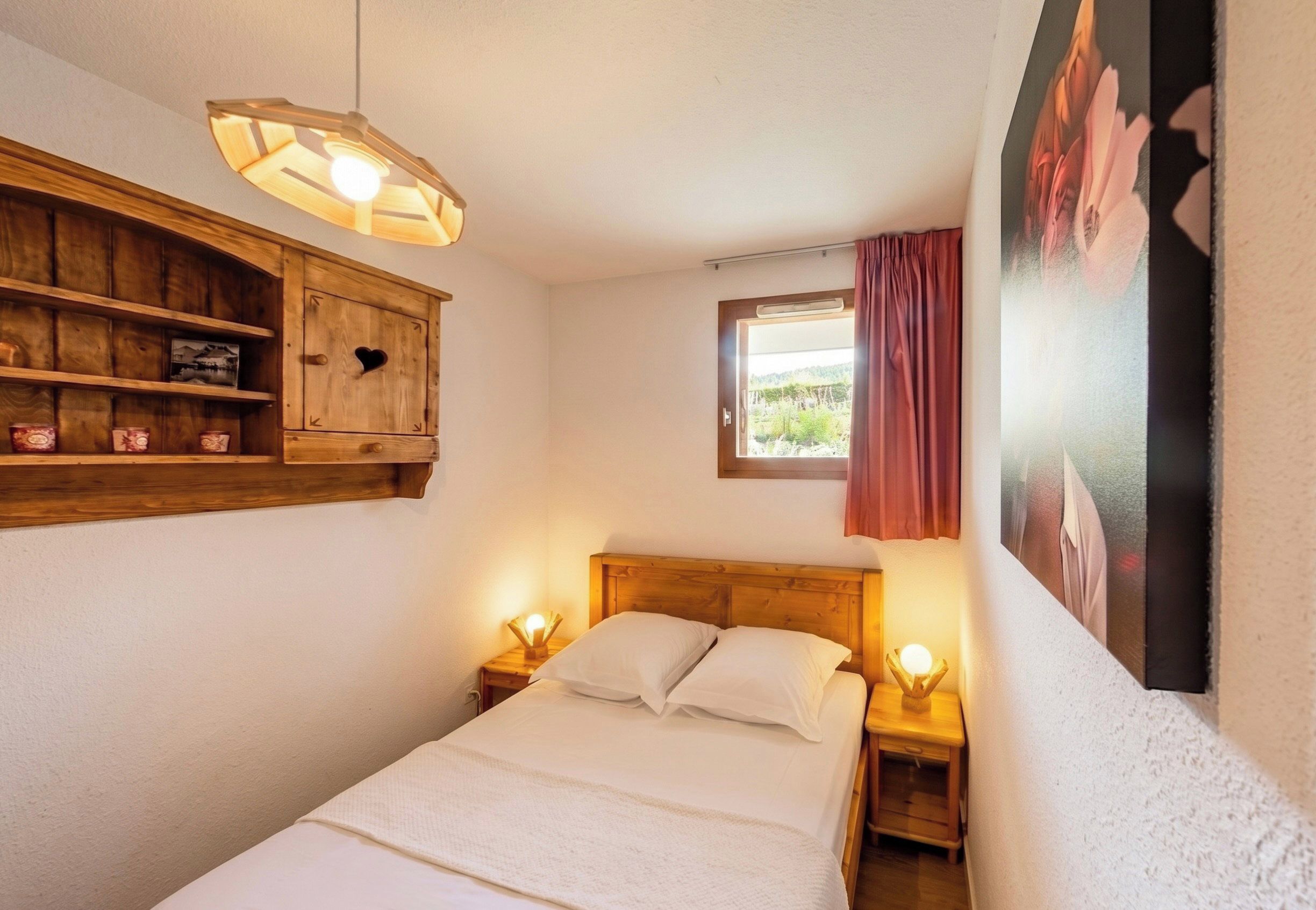 Een slaapkamer met een houten hoofdbord, een nachtkastje met een lamp, een houten kastje met een hartvormig uitsparing, en een raam met uitzicht op groen.