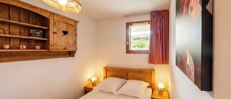 Een slaapkamer met een houten hoofdbord, een nachtkastje met een lamp, een houten kastje met een hartvormig uitsparing, en een raam met uitzicht op groen.