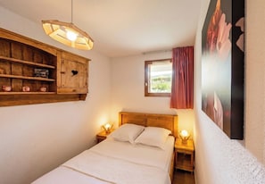 A bedroom with a wooden headboard, a bedside table with a lamp, a wooden cabinet with a heart-shaped cutout, and a window with a view of greenery. - CASA-Amélie terrasse, vue & parking (Vars)