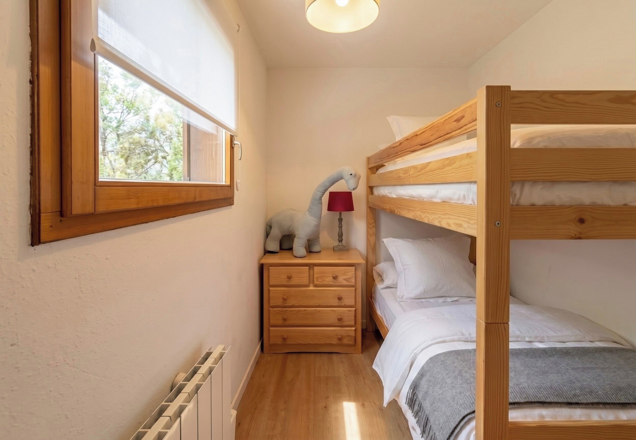 Habitación pequeña con literas, una mesita de noche con lámpara y ventana con vista a árboles.