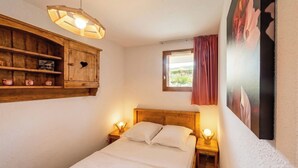A bedroom with a wooden headboard, a bedside table with a lamp, a window with a view, and a wall-mounted cabinet with decorative items. - CASA-Amélie terrasse, vue & parking (Vars)