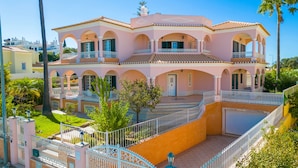 Een roze, twee verdiepingen tellend huis met een balkon, een overdekte patio en een omheinde tuin.