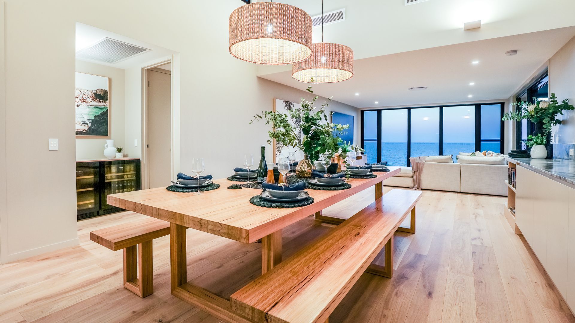 Un comedor moderno con una mesa de madera preparada para seis personas, rodeado de amplios ventanales con vistas al mar.