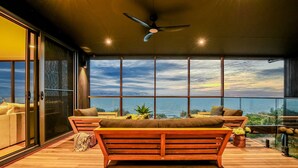 Ein geräumiger Balkon mit einer hölzernen Bank und Kissen, große Fenster mit Blick aufs Meer und ein Deckenventilator.