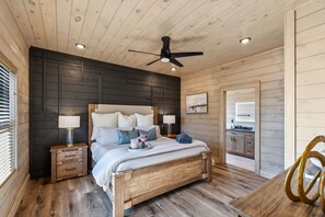 Ein Schlafzimmer mit Holzboden, einem Bett mit Kissen, einem Nachttisch mit Lampe und Blick in die Küche.