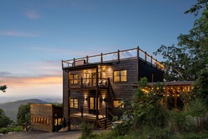 Eine zweistöckige Holzhütte mit Balkon und überdachter Veranda, umgeben von viel Grün und mit Blick auf eine gebirgige Landschaft.