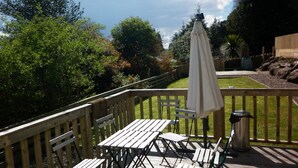 En træterrasse med picnicborde og en parasol, omgivet af frodig vegetation og en stenvæg.
