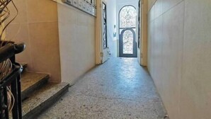 Un couloir étroit, dont le sol est recouvert de carreaux, mène à une porte ornée d’un vitrail décoratif.