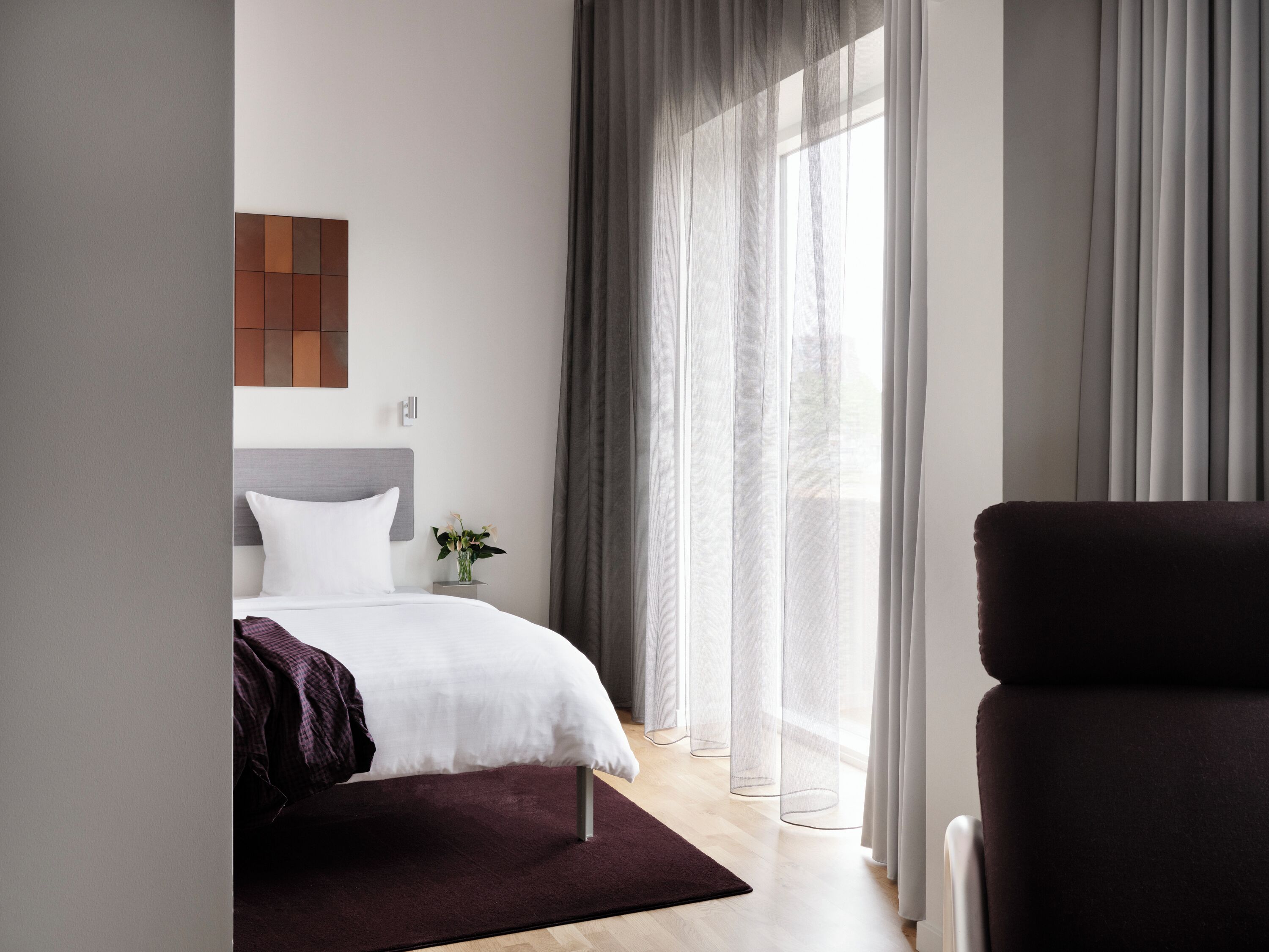 Habitació d'hotel moderna amb un llit, una cadira, una taula petita amb un vas i una finestra amb cortines translúcides.