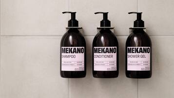 Trois flacons de produits de soins capillaires de la marque Mekano : un shampooing, un après-shampooing et un gel douche, disposés sur un mur carrelé.