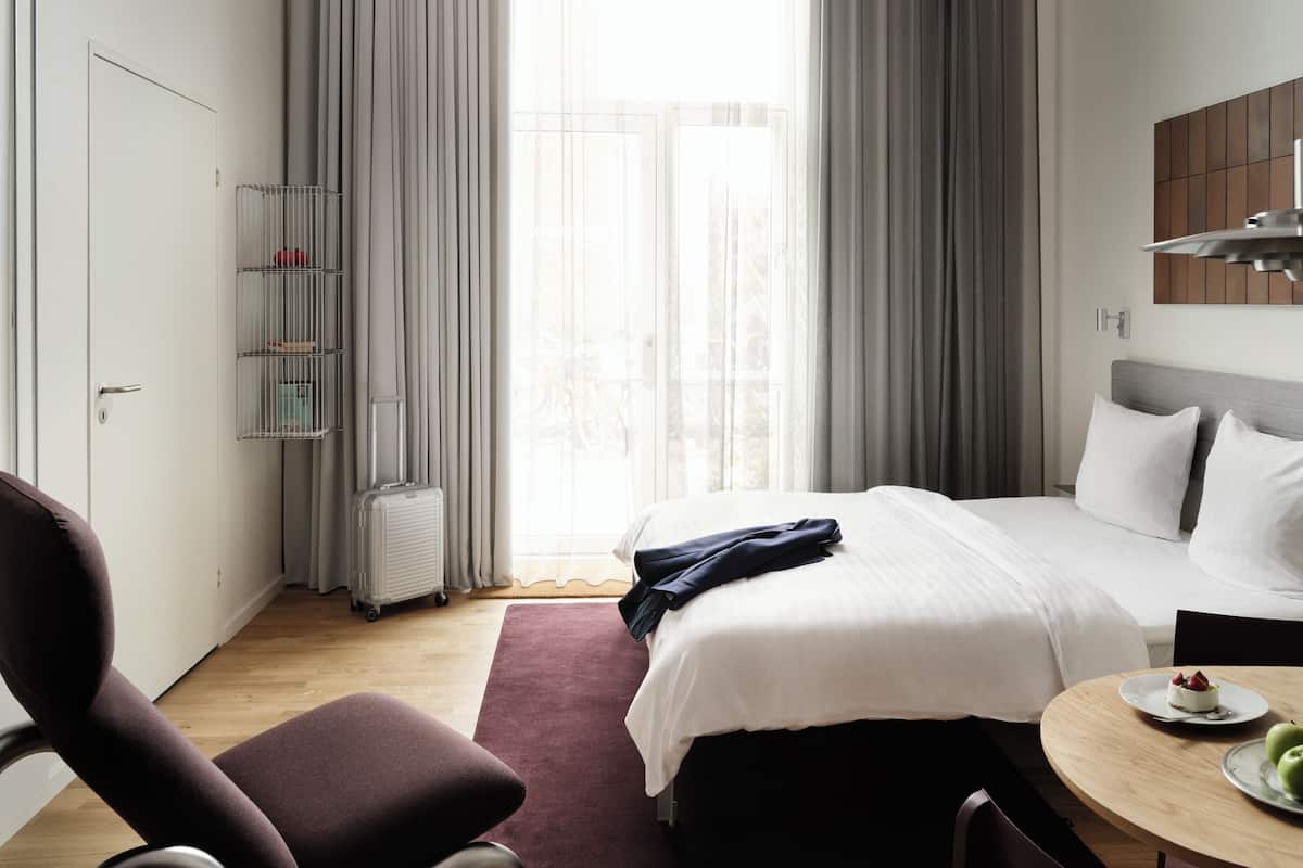 Une chambre d’hôtel moderne avec un lit, une chaise, une petite table sur laquelle se trouve une assiette de nourriture, et une fenêtre avec des rideaux.