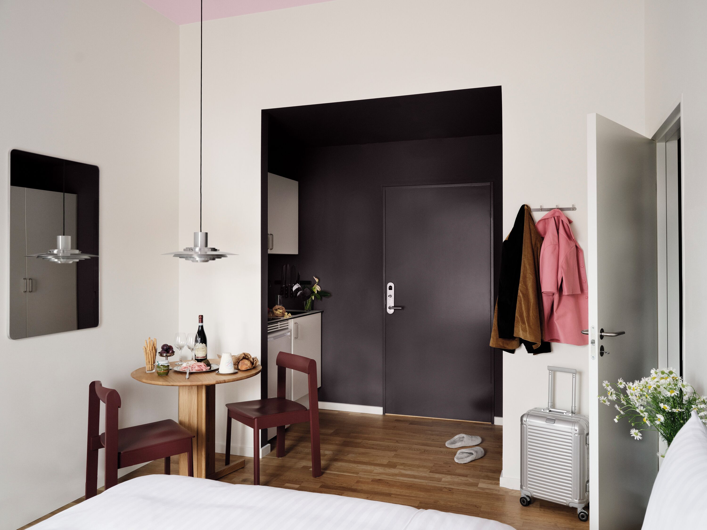 A hotel room with a bed, a desk with a chair, a small table with food and drinks, a suitcase, and a door leading to a bathroom.