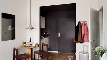A hotel room with a bed, a desk with a chair, a small table with food and drinks, a suitcase, and a door leading to a bathroom.
