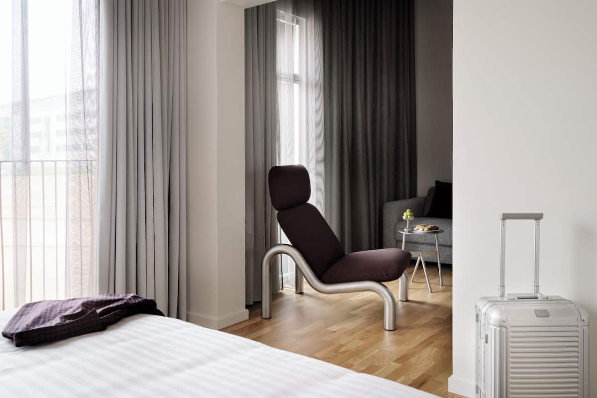 Une chambre d’hôtel avec un lit, une chaise, une petite table sur laquelle se trouve un vase, et une valise.