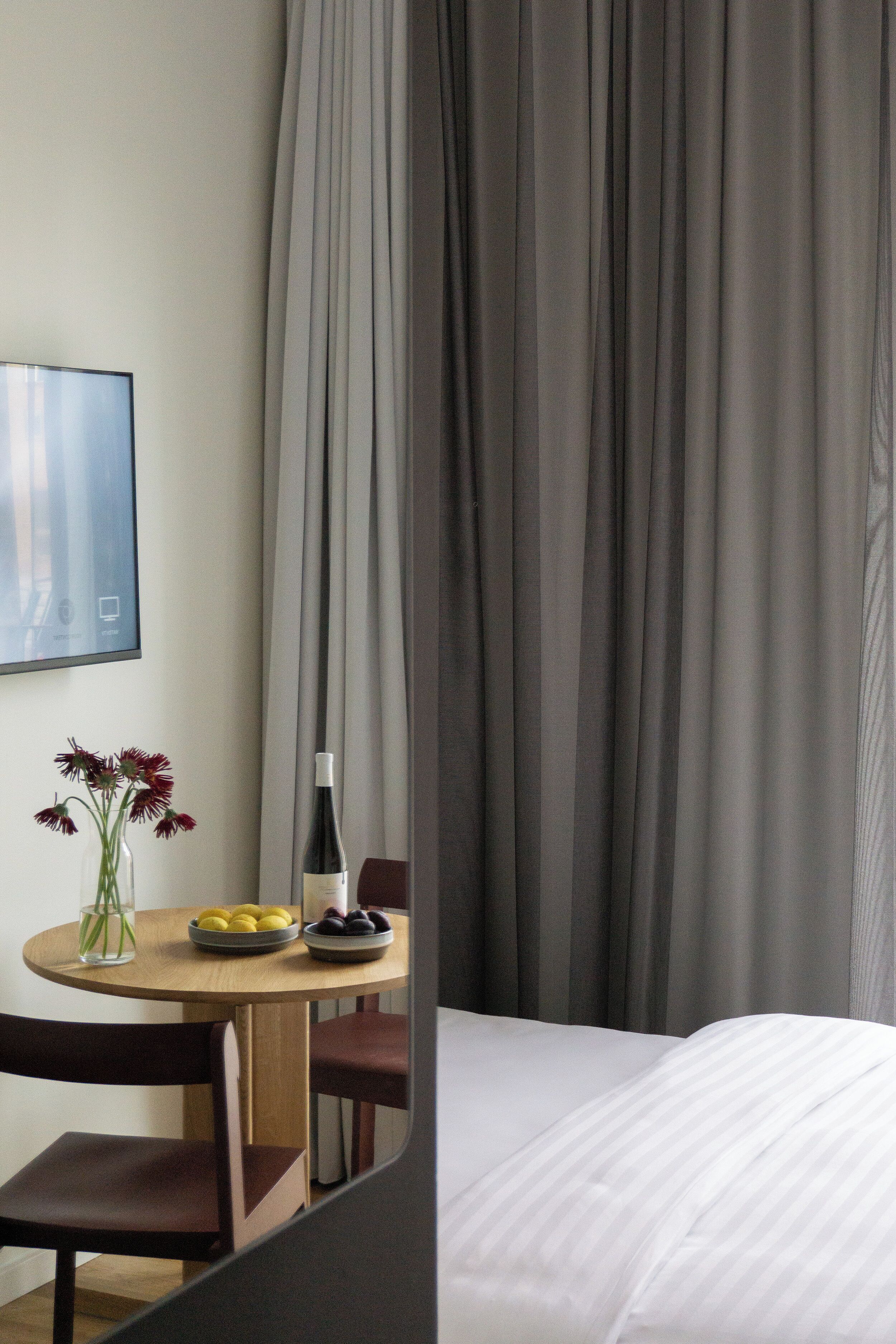 A hotel room with a bed, a round wooden table with a vase of flowers, a plate of fruit, and a bottle of wine.