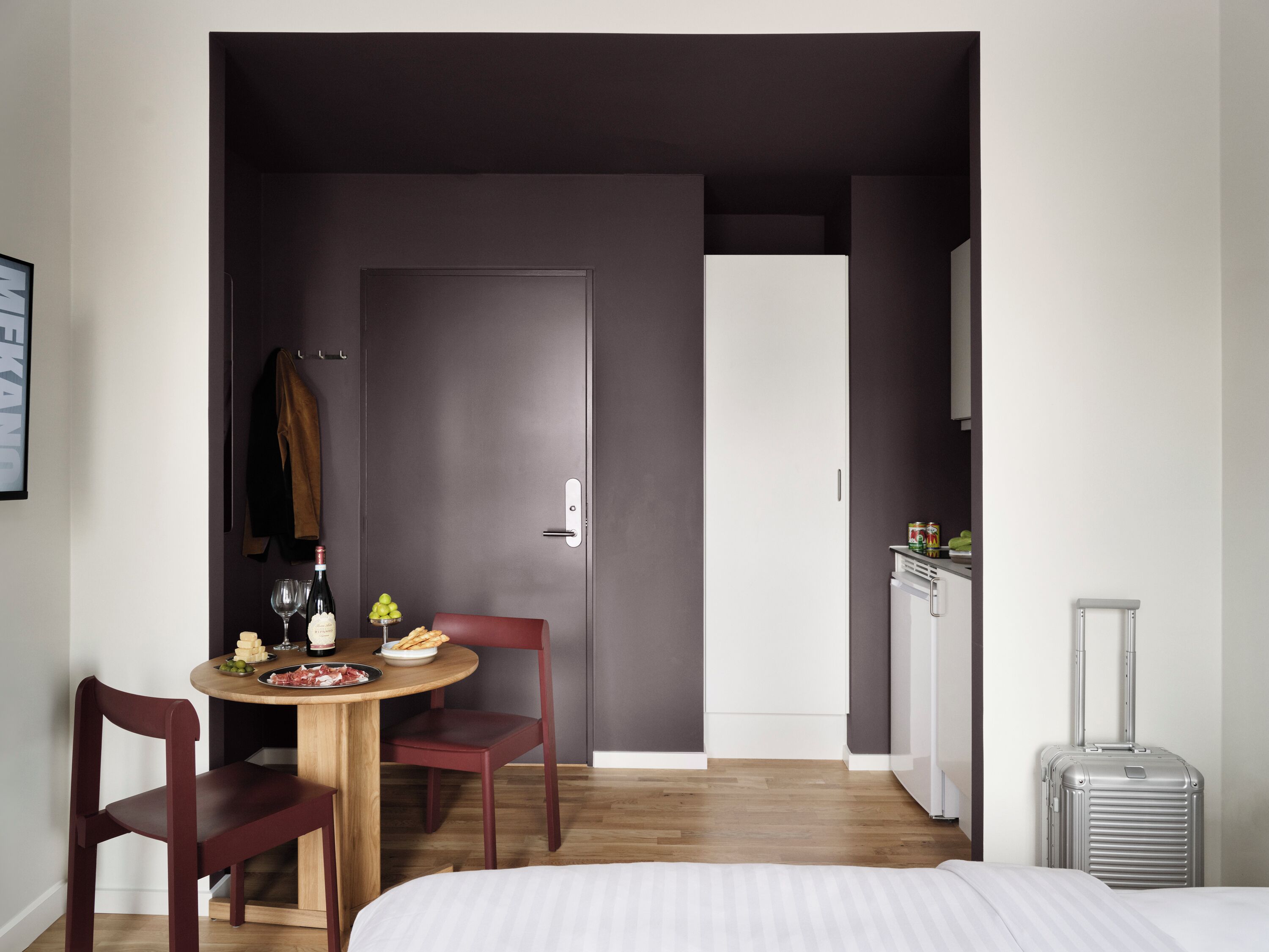Habitació d'hotel senzilla amb llit, taula petita de dinar amb cadires, barra minibar i maleta.