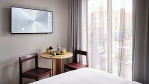 Ein Hotelzimmer mit einem Bett, einem an der Wand befestigten Fernseher, einem runden Holztisch mit Frühstücksangeboten, zwei Stühlen und einem großen Fenster mit Vorhängen.