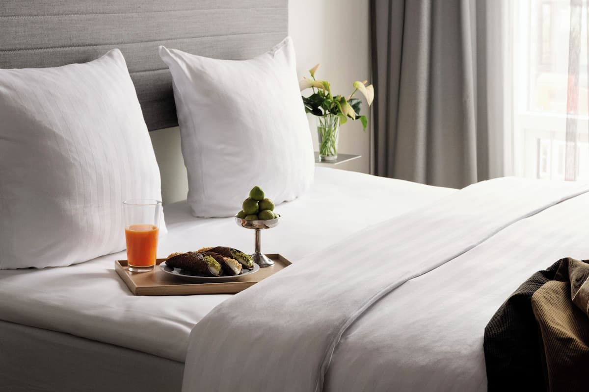 Un lit bien fait, avec du linge de lit blanc, une table de chevet sur laquelle se trouvent un verre de jus d’orange et une assiette de nourriture, et une vue sur l’extérieur par une fenêtre ornée de rideaux.