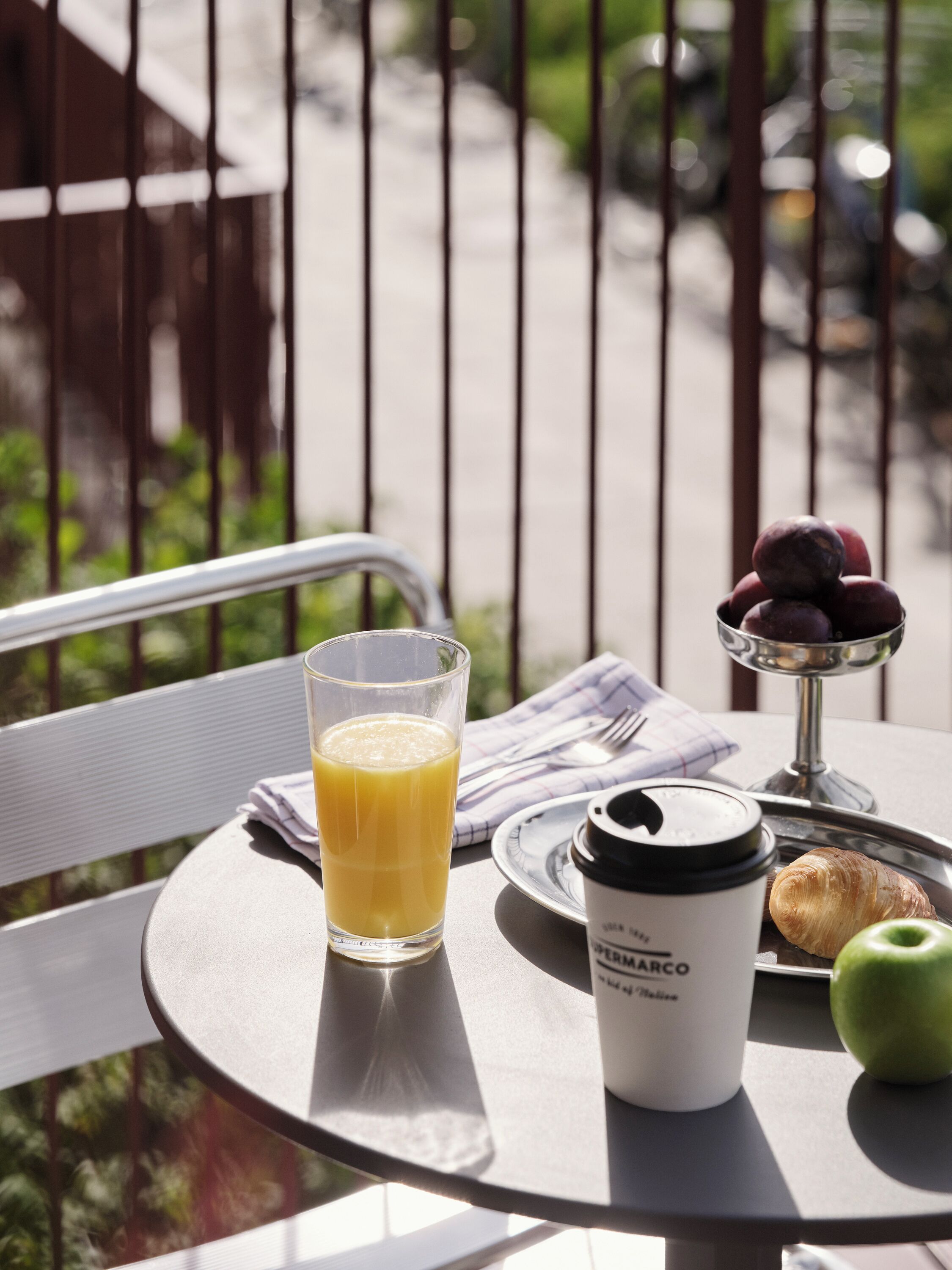A table with a glass of orange juice, a coffee cup, a plate with plums, a croissant, and an apple.