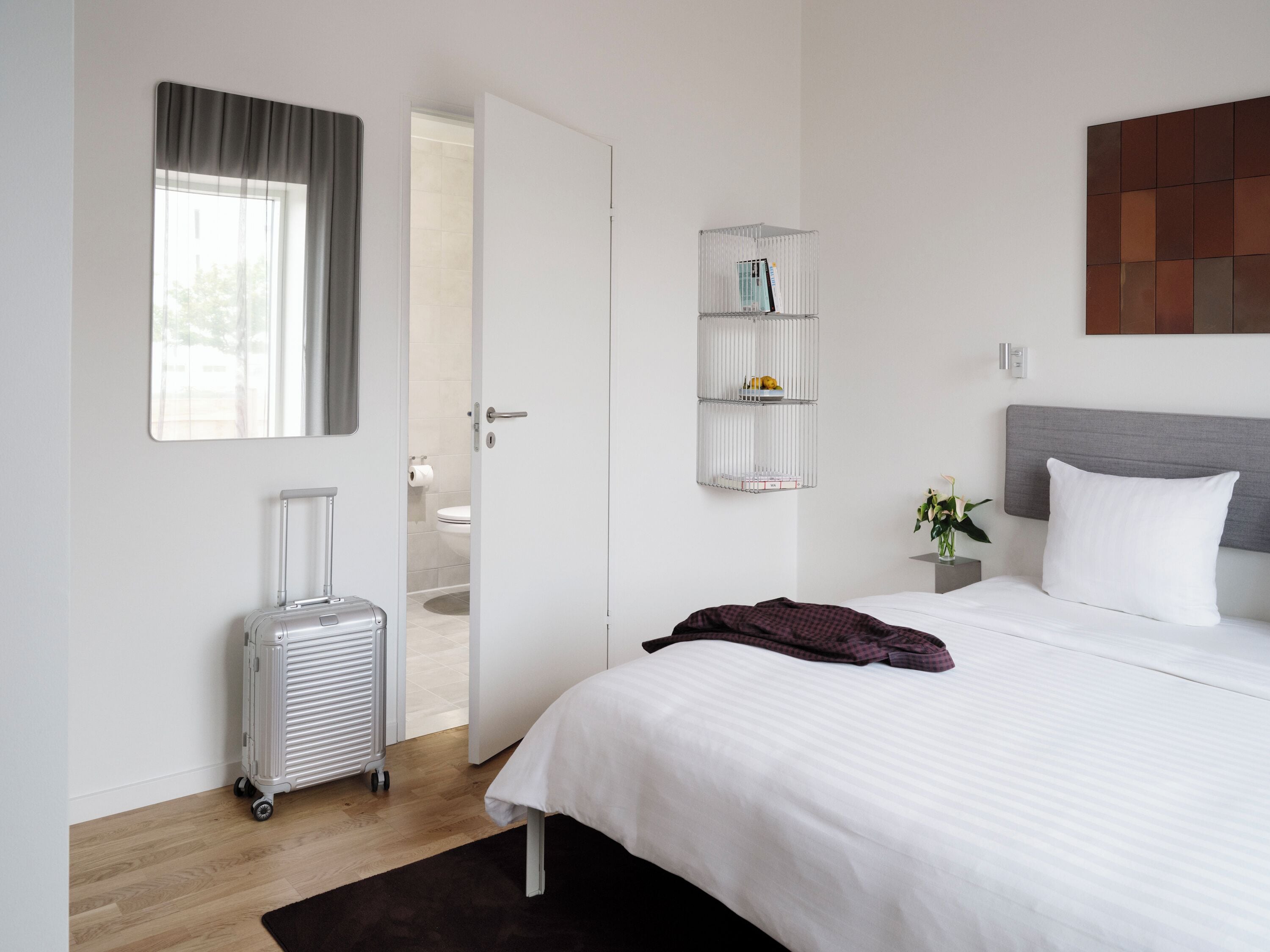 A hotel room with a bed, a suitcase, a bathroom visible through an open door, and a wall-mounted shelf with books.