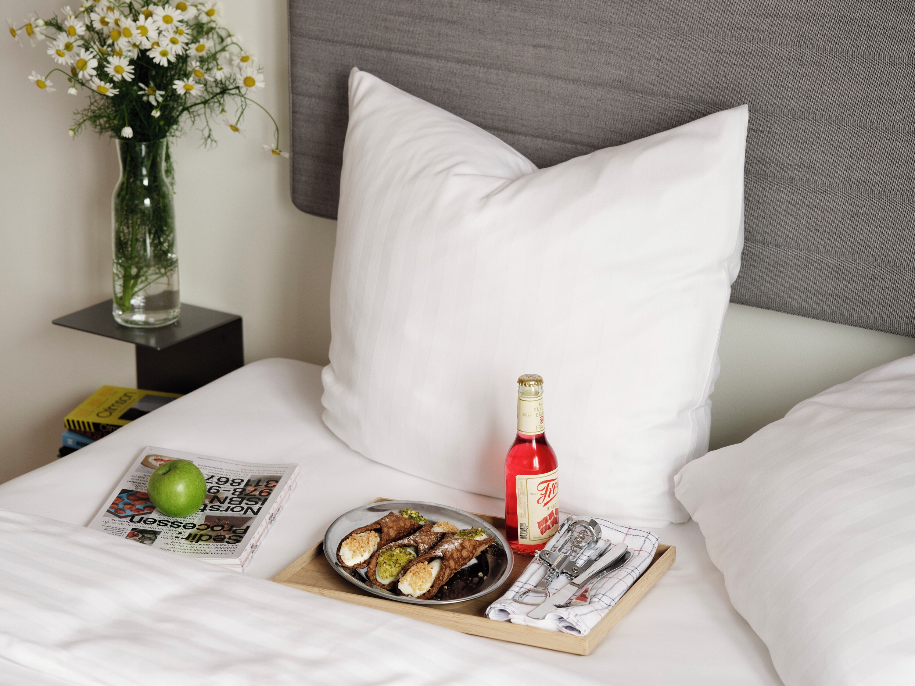 Una cama bien hecha con sábanas blancas, una botella de vino tinto, un plato de sushi, una manzana verde y una revista en una mesita de noche.