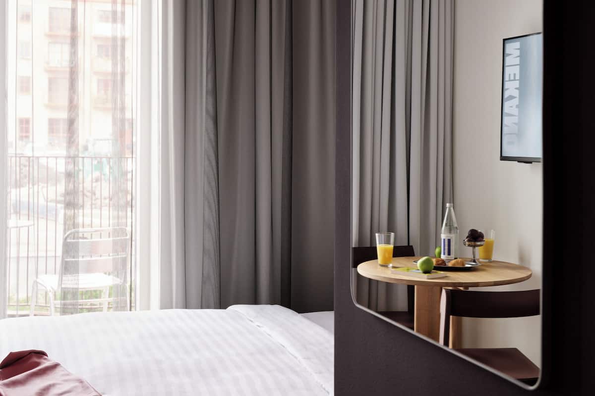 Une chambre d’hôtel avec un lit, une table ronde sur laquelle sont disposés des aliments pour le petit-déjeuner, une chaise et une grande fenêtre avec des rideaux.