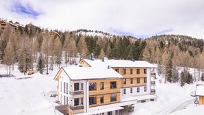 Um hotel com várias andares, fachada de madeira e varandas, cercado por árvores cobertas de neve e um cenário montanhoso.