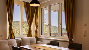 Uma sala de jantar com mesa e cadeiras de madeira, um sofá e janelas amplas com cortinas.