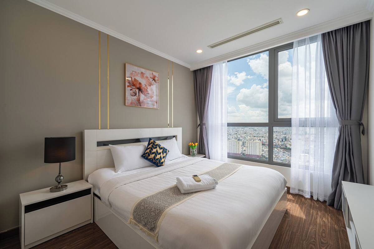A hotel room with a large bed, a nightstand with a lamp, a city view through the window, and a painting on the wall.
