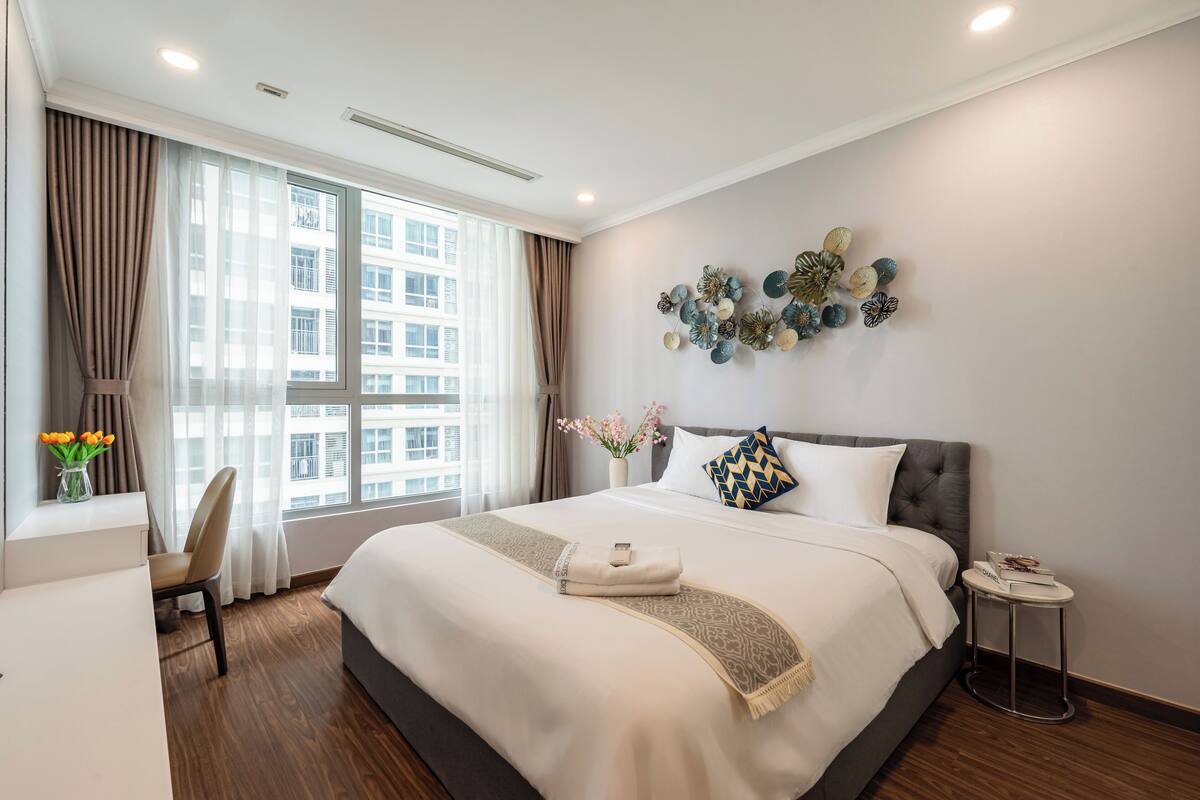 A hotel room with a large bed, a desk, a chair, a small side table, and a window with curtains.
