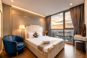 Une chambre d’hôtel avec un grand lit, un fauteuil bleu, une table de chevet avec une lampe, et une vue sur la ville par une grande fenêtre.