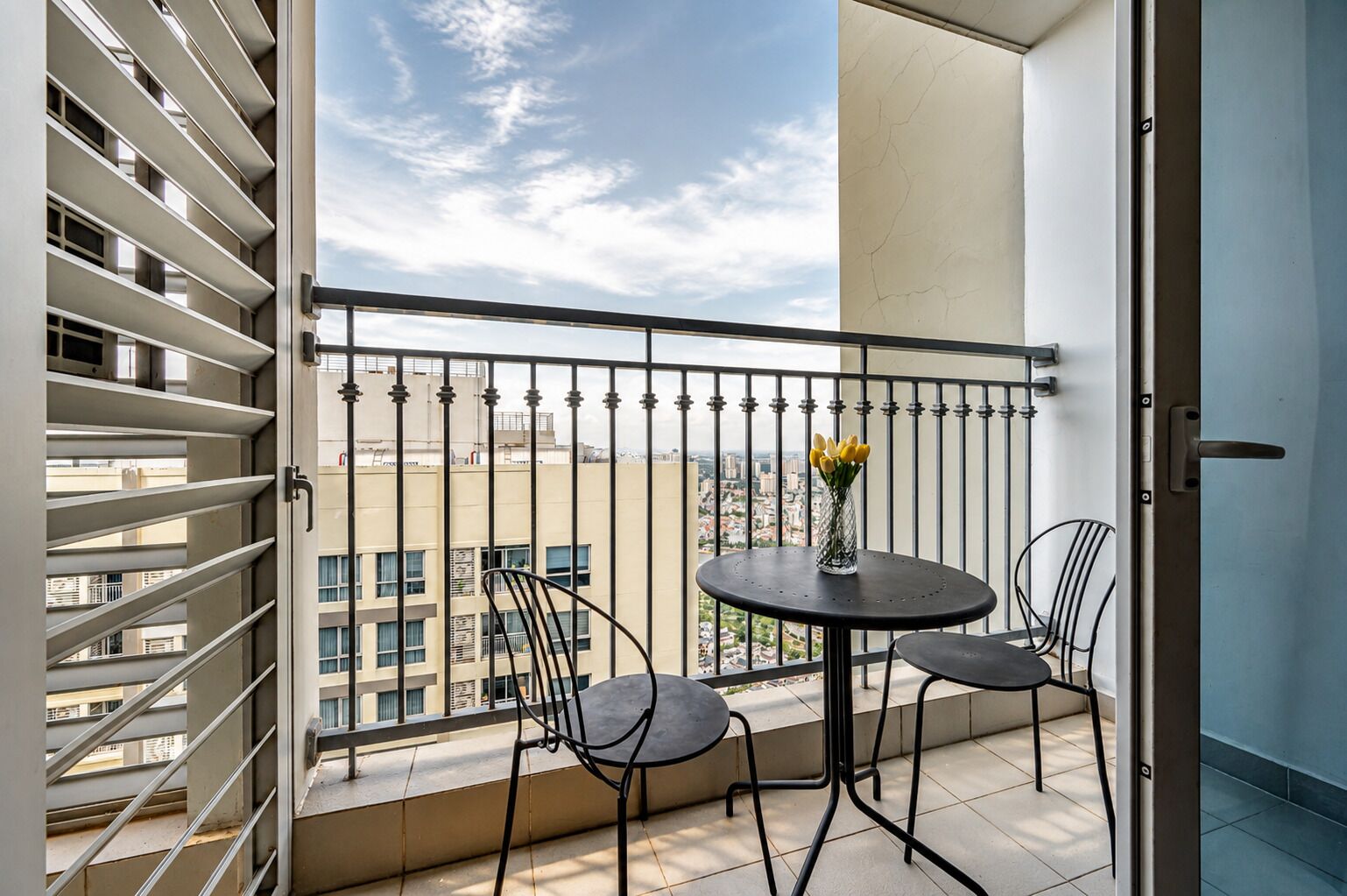 A balcony with a table and chairs, a vase with flowers, and a cityscape view.