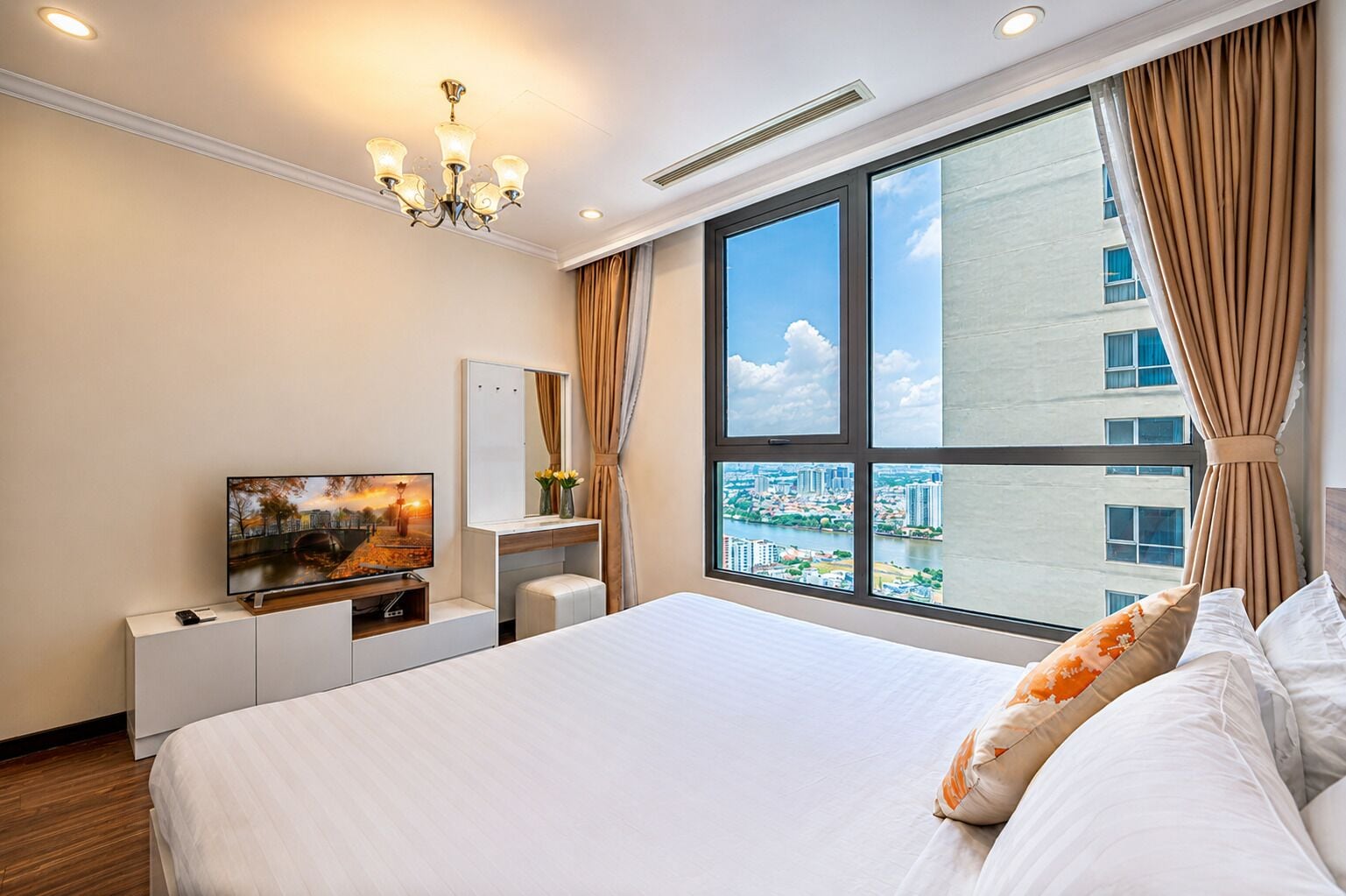 A hotel room with a large bed, a TV, a desk with a mirror, and a view of the city through large windows.