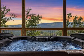 Un jacuzzi con vistas a montañas y a un atardecer, todo visible a través de amplios ventanales.