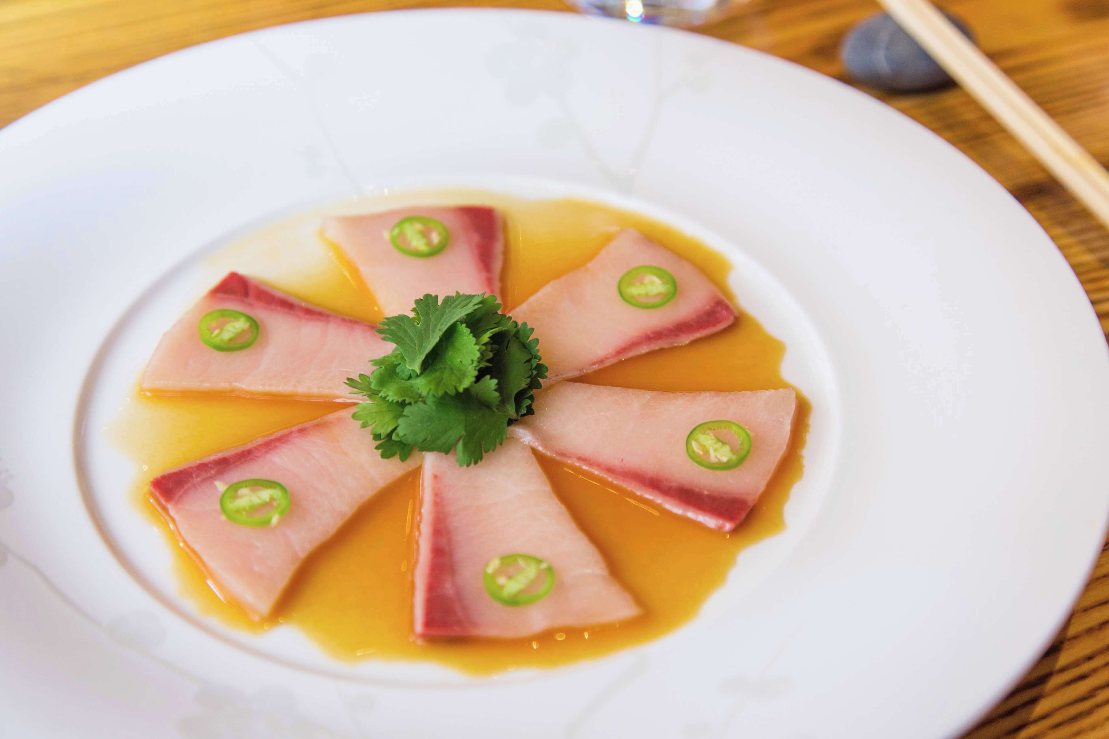 A white plate with a dish of thinly sliced meat arranged in a circular pattern, garnished with cilantro and green chili slices.