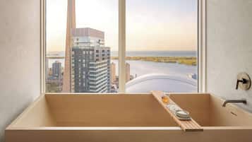 Un baño moderno con una bañera grande, un taburete de madera y vistas a un paisaje urbano que incluye una torre y edificios.