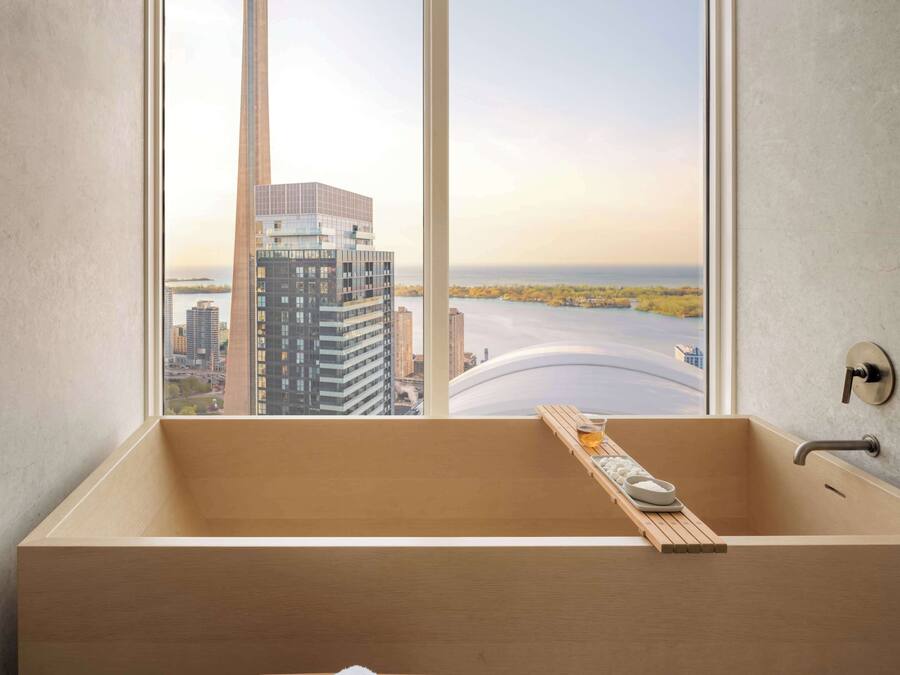 Un baño moderno con una bañera grande, un taburete de madera y vistas a un paisaje urbano que incluye una torre y edificios.