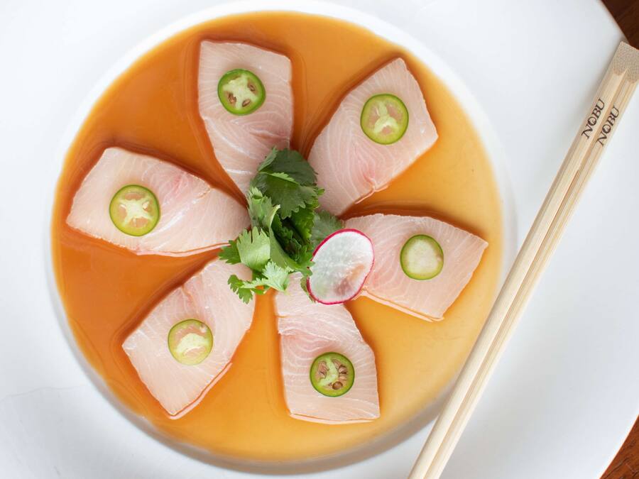 A bowl of sashimi arranged in a circular pattern with a clear broth, garnished with radish slices and jalapeño peppers.