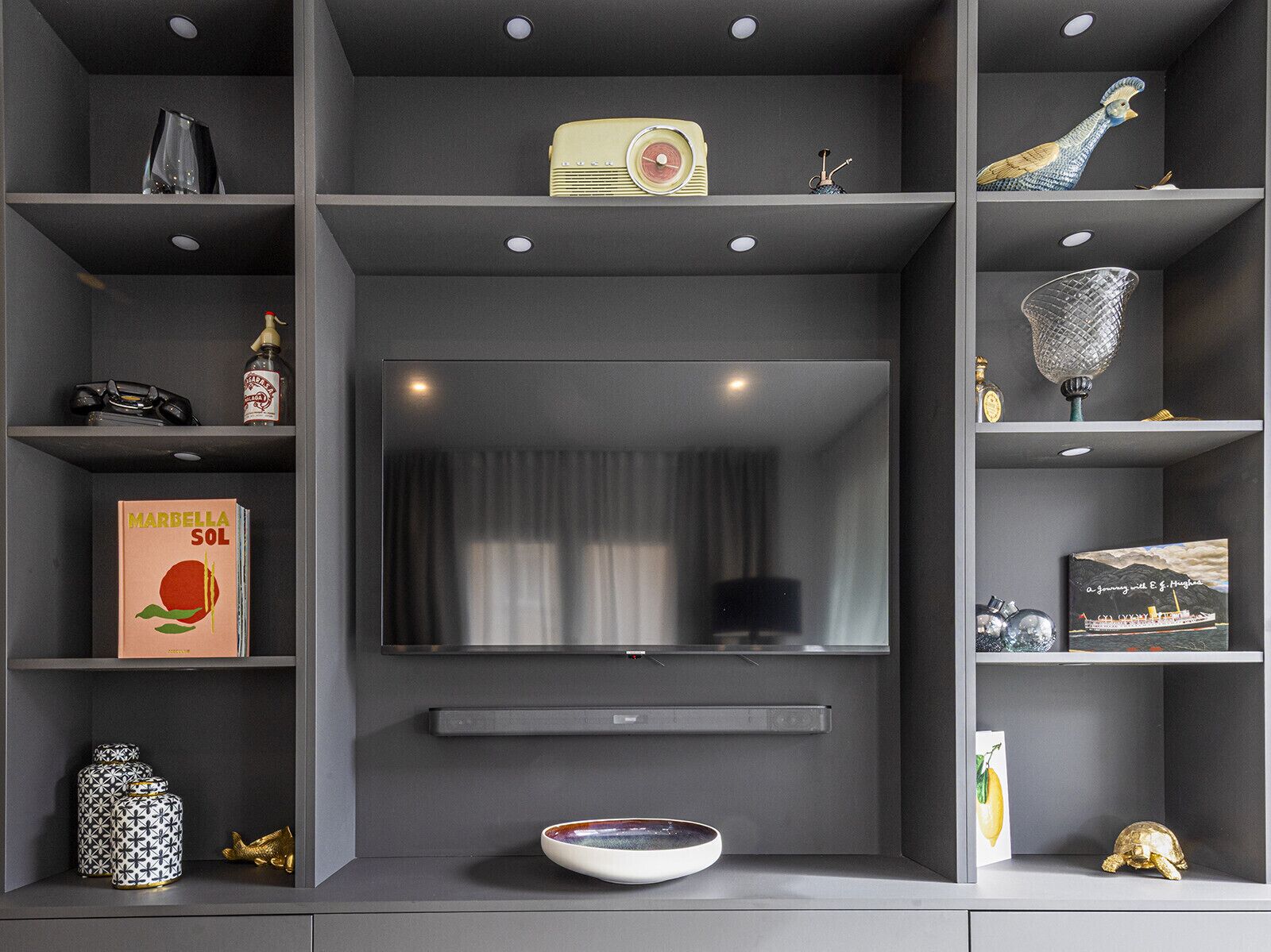 Un salon moderne doté d’un grand téléviseur à écran plat fixé au mur, entouré d’étagères où sont disposés divers objets décoratifs, et un bol posé sur le sol.