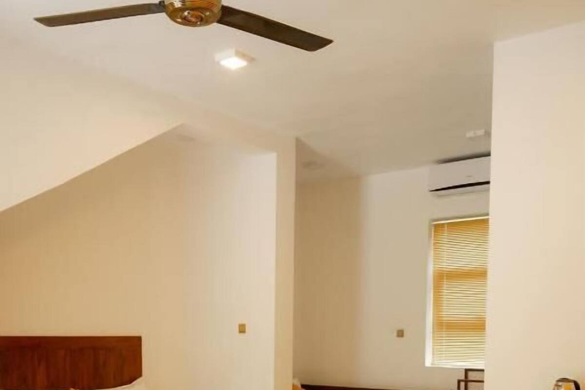 A hotel room with two beds, a ceiling fan, a wooden headboard, a small table with a kettle, and a window with blinds.