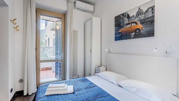 Ein Hotelzimmer mit Bett, Fenster mit Vorhängen, Heizkörper, einem kleinen Nachttisch und einer Wand-Klimaanlage.