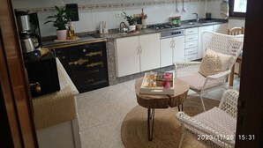 Een moderne keuken met witte kasten, een gasfornuis en een koffieapparaat. Er staat een rieten stoel met kussen en een klein houten tafeltje met een dienblad.
