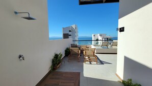 A balcony with wicker chairs and a glass table overlooking a modern building and the sea.