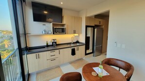 A modern kitchen with a flat-screen TV, a microwave, a coffee maker, and a refrigerator. There is a dining table with chairs and a view of the outside.