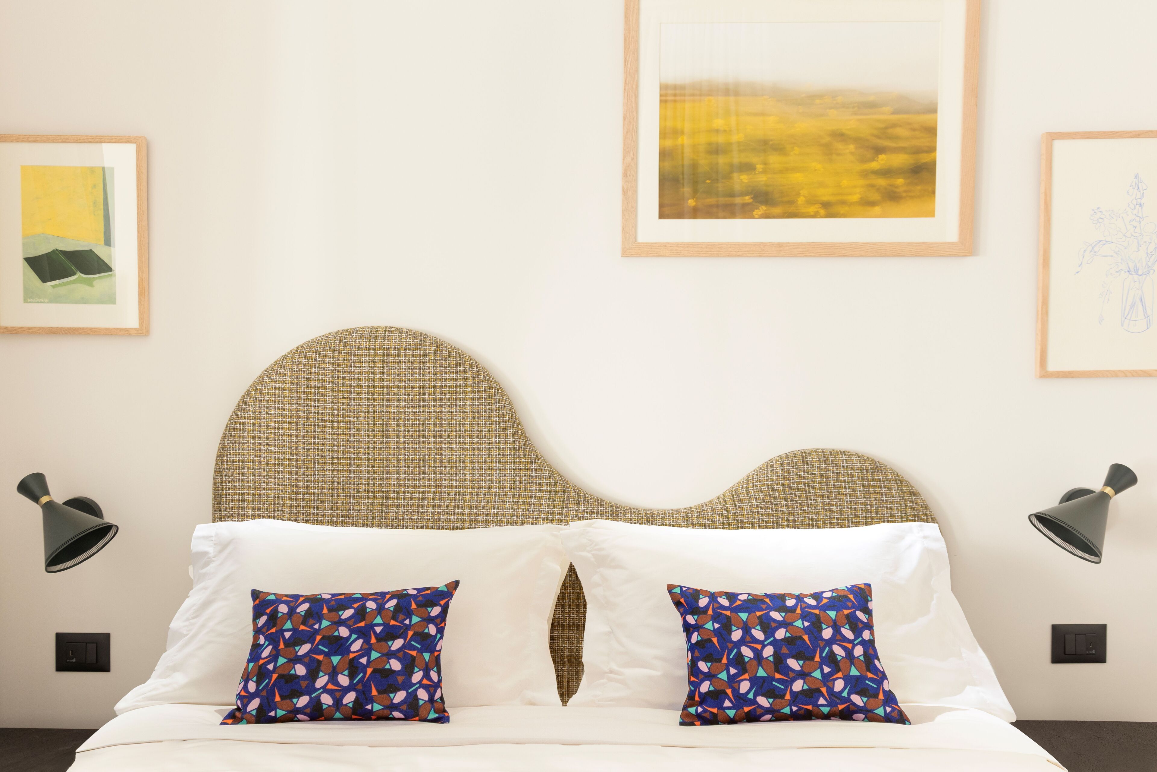 A neatly made bed with white linens and two patterned pillows, a woven headboard, and framed artwork on the walls.