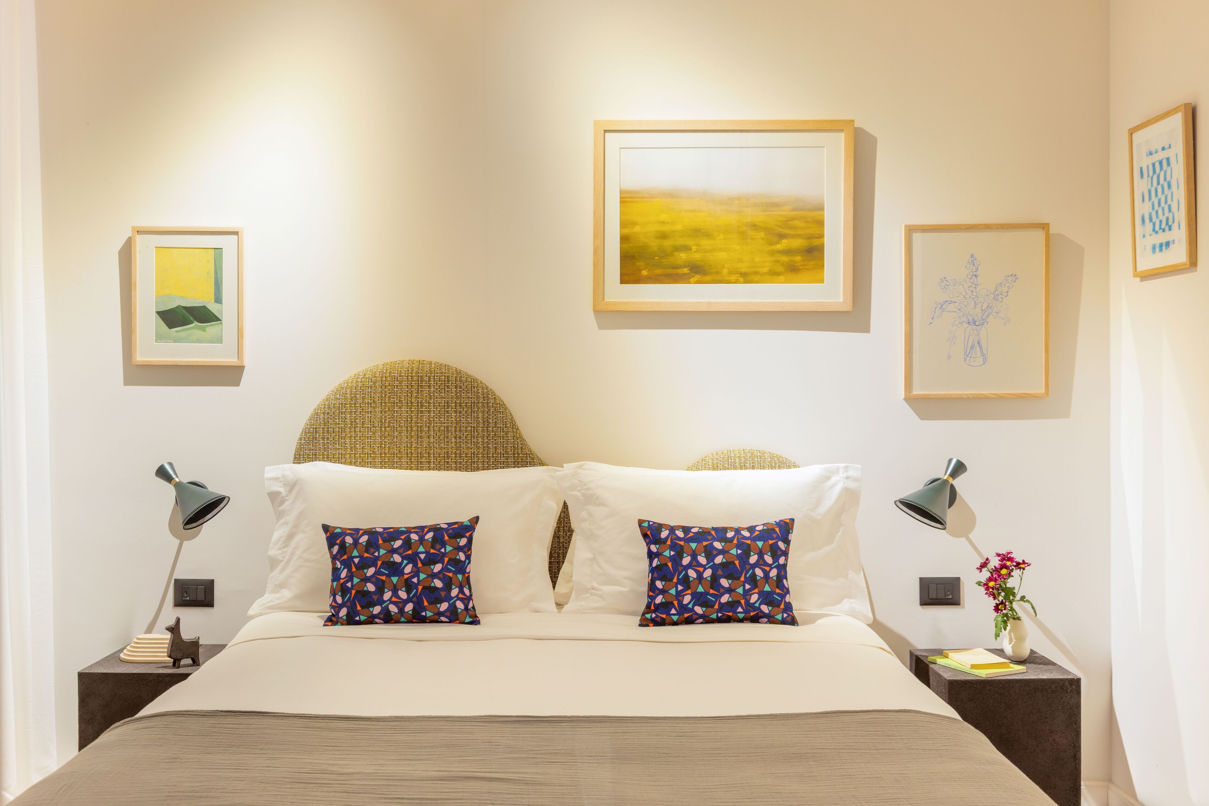 A neatly arranged bedroom with a bed, two pillows, a nightstand with a lamp, and framed artwork on the wall.