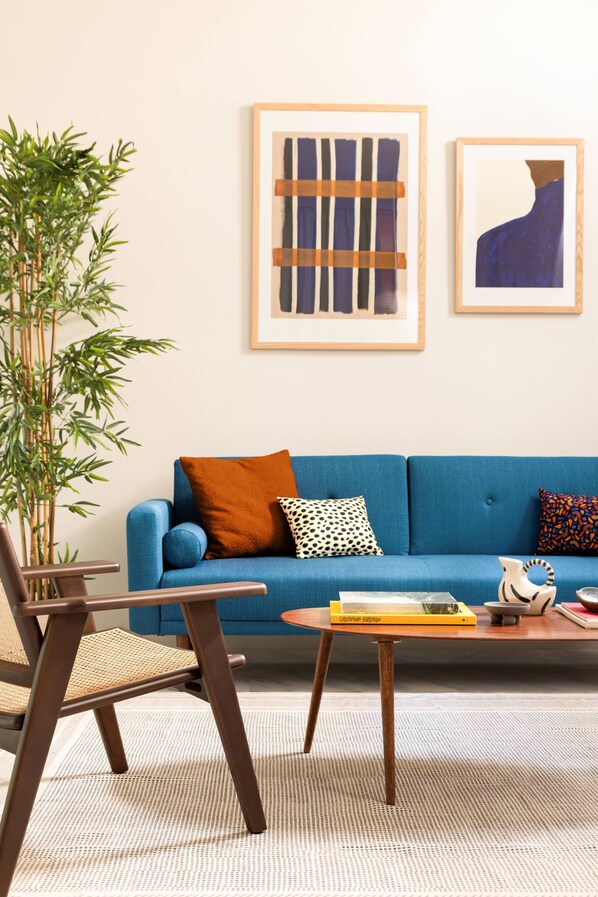 A living room with a blue sofa, a wooden chair, a small table with books, and two framed abstract paintings on the wall. - Silk n Cotton (Athens)