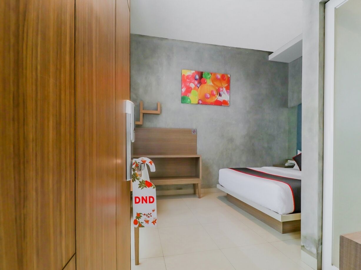 Ein kleines Hotelzimmer mit einem Bett, einer hölzernen Sitzbank, einem kleinen Tisch und einem an der Wand befestigten Kunstwerk.